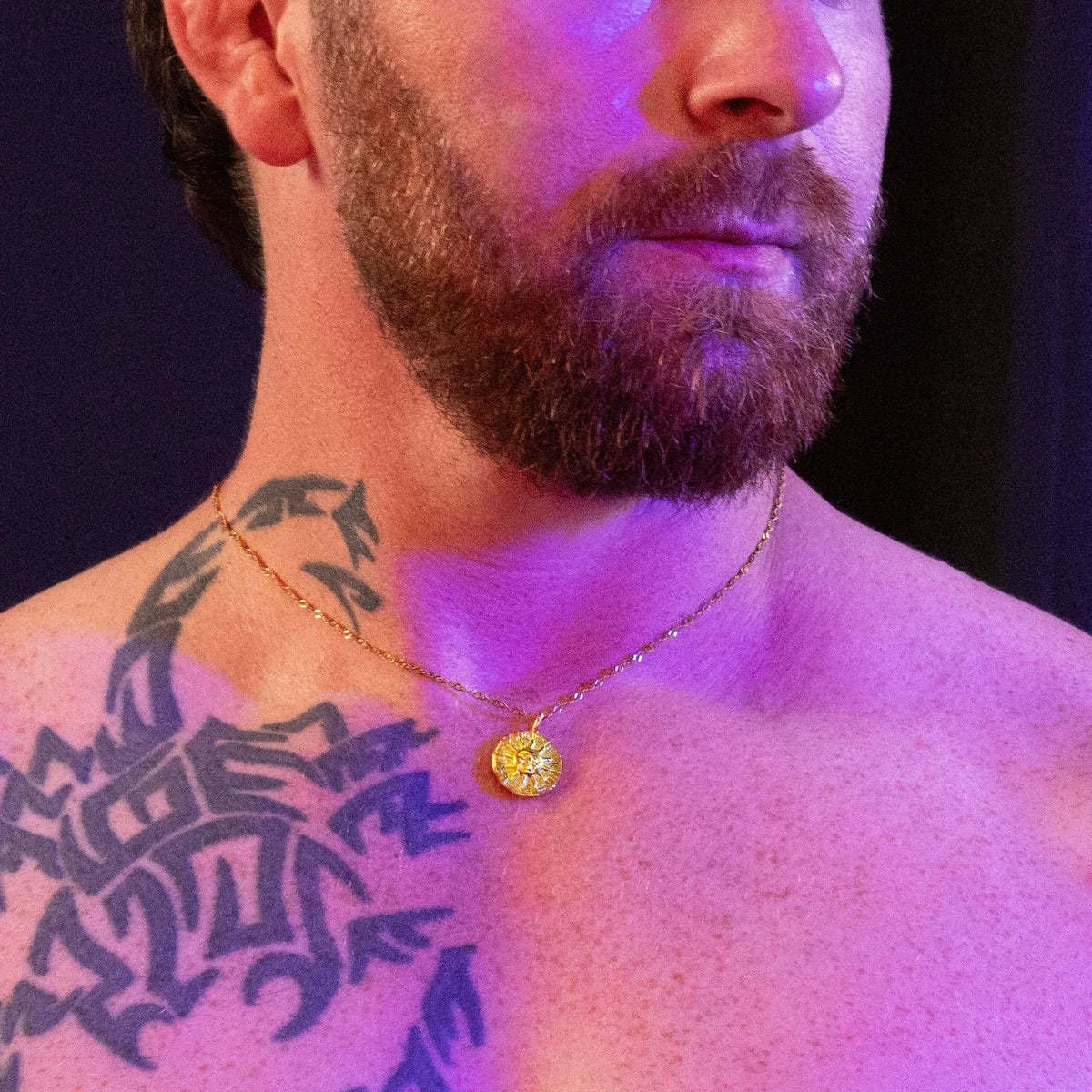 Gold medallion necklace worn against purple-lit skin with a dark tribal tattoo visible.