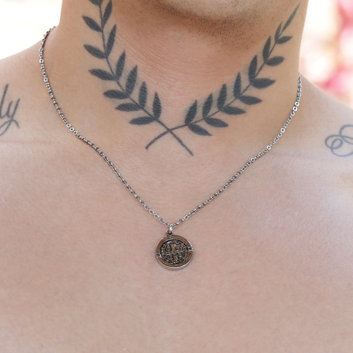 Silver necklace with a circular pendant worn over a neck tattoo of crossed laurel branches.