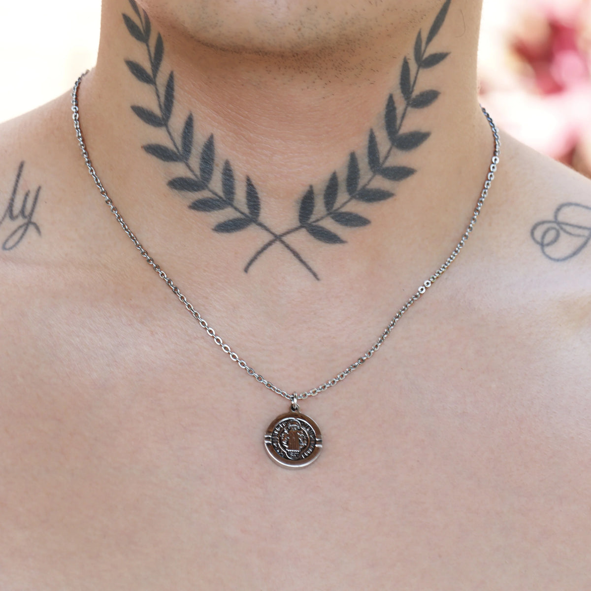 Tattoo of crossed laurel branches on a person’s neck, with a silver necklace below it.