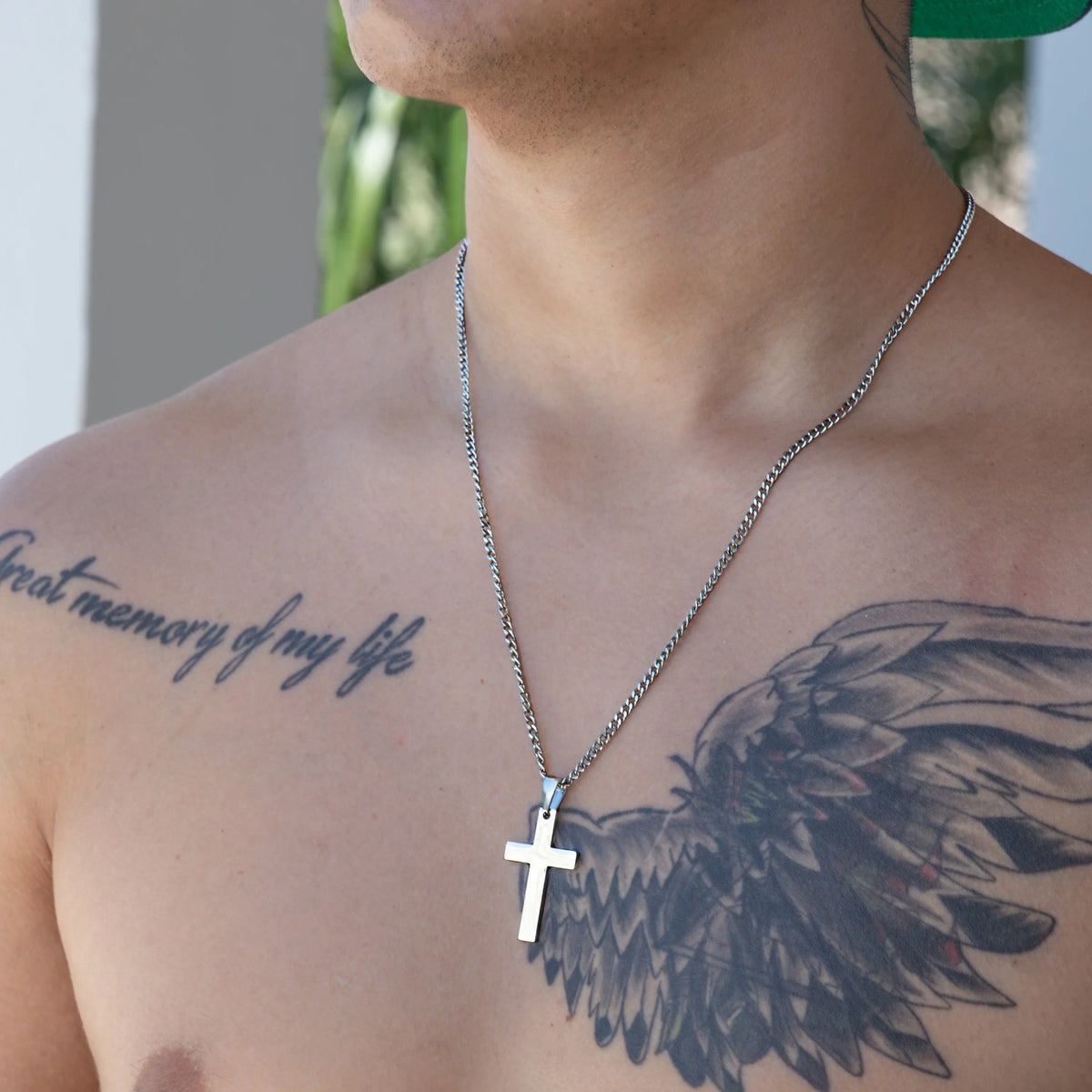 Silver cross pendant necklace worn on a person’s chest with visible tattoos.