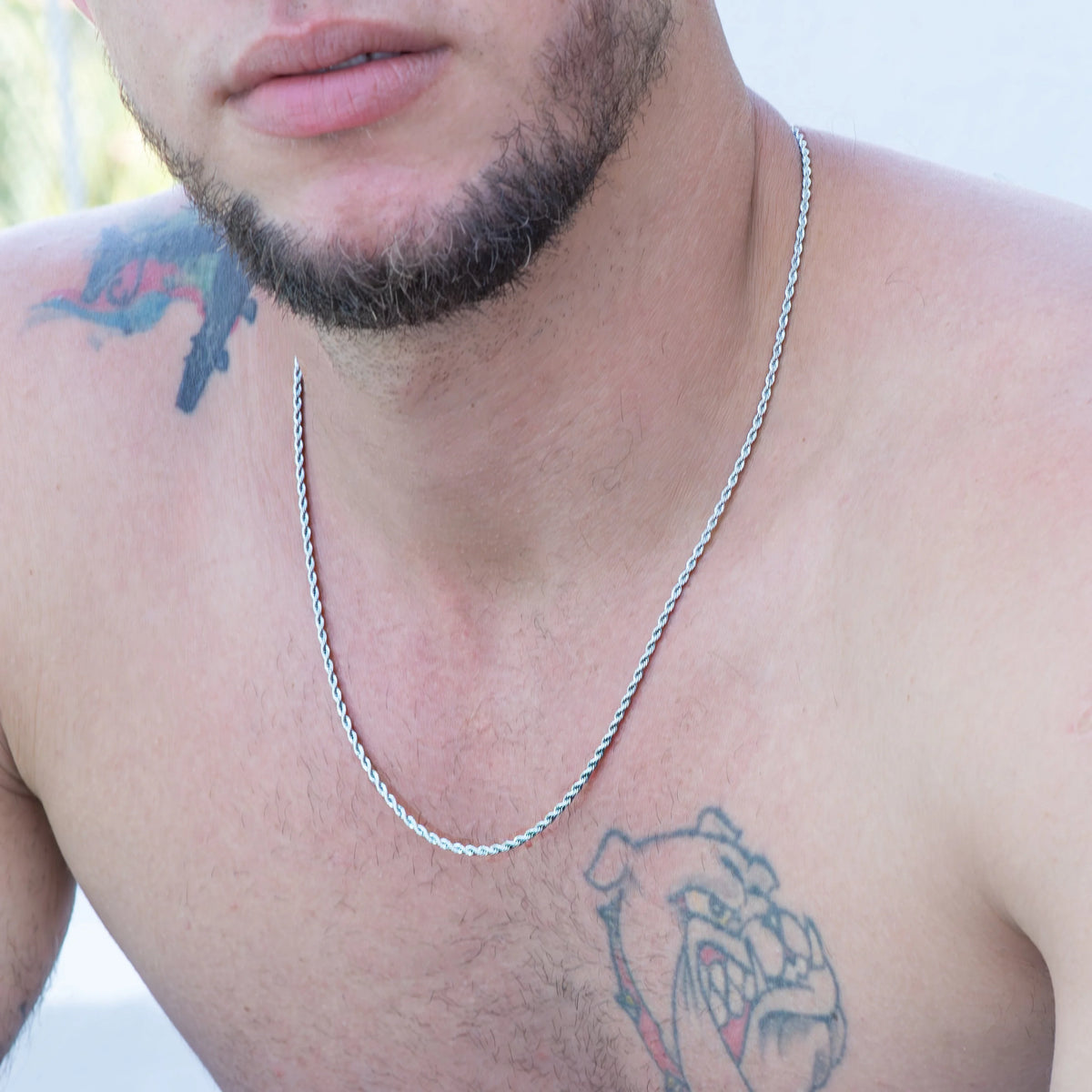 Close-up of a man’s upper chest and lower face, showing tattoos and a silver necklace.