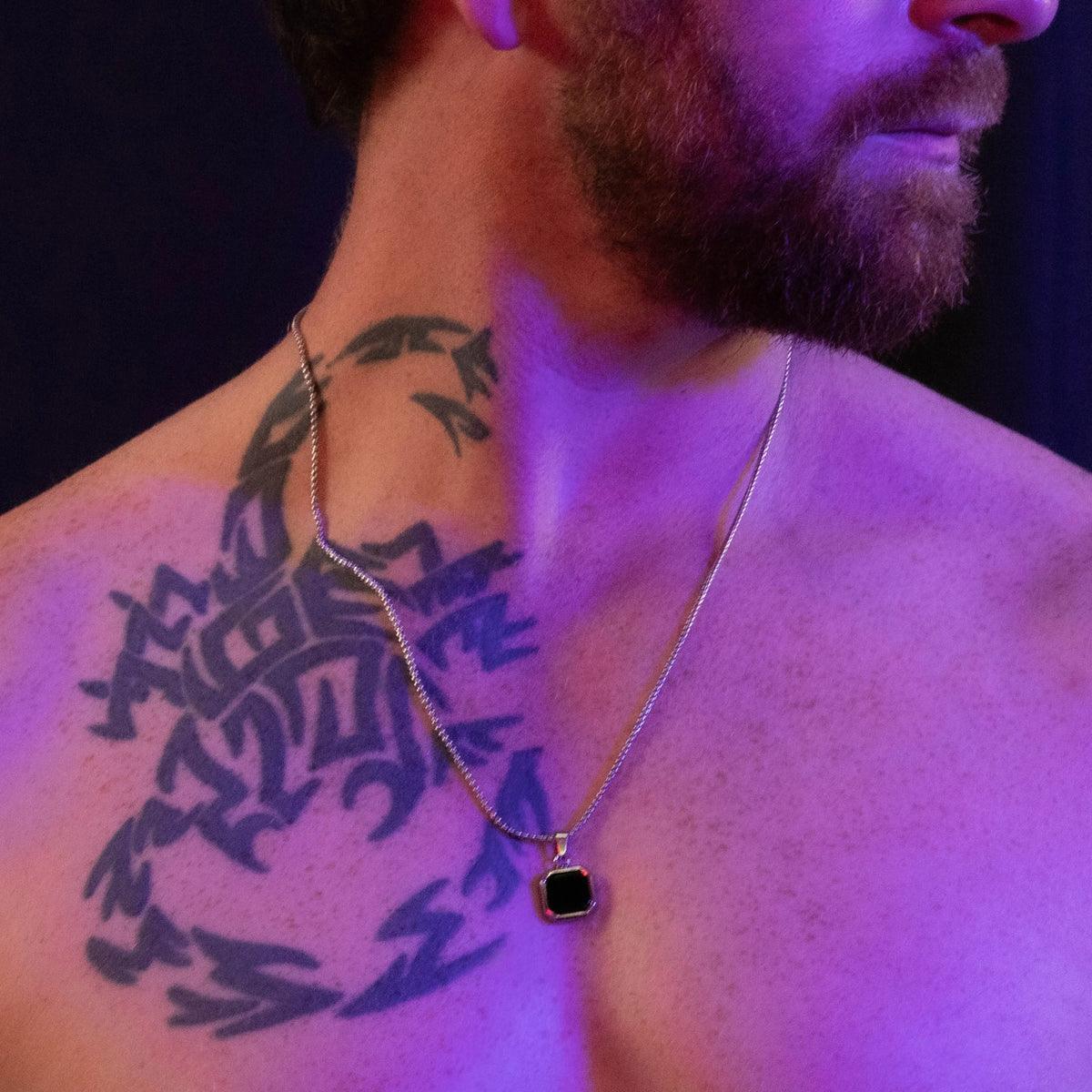 Black pendant necklace on a silver chain against tattooed skin under purple lighting.